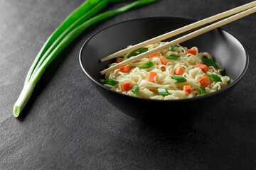 Instant noodles, cooked with vegetables and green onion, in gray bowl, chopsticks, on dark background, selective focus.