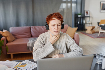 Horizontal shot of mature woman suffering flu with sore throat wearing headphones sitting at desk...