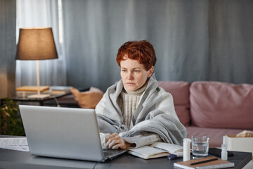 Portrait of mature woman having flu wearing warm clothes sitting at desk in living room at home working on laptop