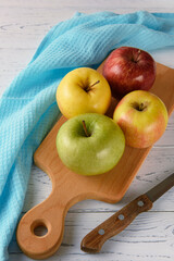 Multi-colored apples lie on a wooden board on a white wooden background. Nearby lie a blue napkin and a knife.