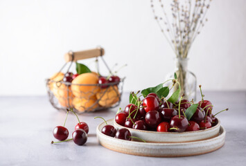 Fresh organic cherries in a grey ceramic plate on a light grey background. In the background there is a basket with apricots and cherries and a saucer with cherries. Upper angle.