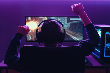 Back view of pro gamer wearing headset celebrating victory in online internet esport tournament, makes yes hands gesture, looking at monitor. Neon coloured dark room. Cyber sport and e-sport concept - obrazy, fototapety, plakaty