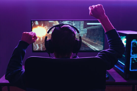 Back View Of Pro Gamer Wearing Headset Celebrating Victory In Online Internet Esport Tournament, Makes Yes Hands Gesture, Looking At Monitor. Neon Coloured Dark Room. Cyber Sport And E-sport Concept