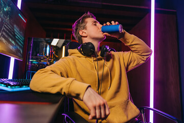 Portrait of young professional cyber sportsman sitting at gaming desk at home and drinking caffeine energy drink to concentrate on online video shooter game at night. Neon coloured room. Cyber sport