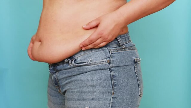 Side view of unrecognizable fat plump overweight woman wearing blue jeans, shaking, lifting belly, squeezing excess flabby stomach on blue background. Body positive, obesity, weight loss, liposuction.