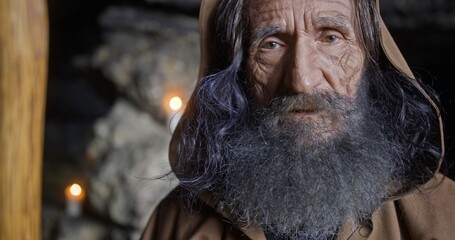 Portrait of senior sage man in cassock and with gray hair standing in rocky cave with candles and looking at camera