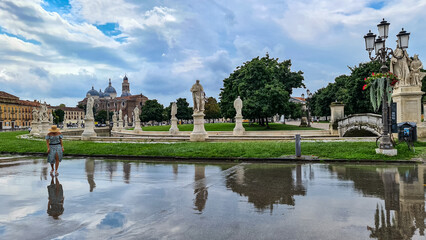 Fototapeta premium Woman walking on the center square after rain. Scenic view after strong rain on Prato della Valle, Abbey of Santa Giustina, city of Padua, Veneto, Italy, Europe. Water on street creates reflection