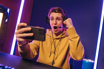 Excited and shocked gamer with headset playing video mobile game online on his smartphone, sitting on gaming chair in neon room. Professional gamer streaming the game, having fun, rejoices in victory - obrazy, fototapety, plakaty