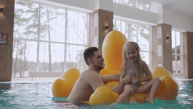 adult man and his little daughter are swimming in pool, child girl is sitting on funny inflatable duck
