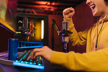 Close up of young exited cyber sportsman shouting and making winner hand gesture, celebrating his winning online shooter video game tournament. Pro gamer streaming his gaming. Site view. Neon color