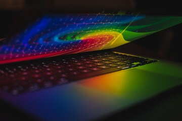 Closeup shot of an open laptop with rainbow light on it and a keyboard reflecting on a screen