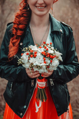 closeup wedding bouquet flowers in hands informal red hair bride in red skirt dress and black leather jacket which stands with happy smile on the spring park background. wedding concept, free space
