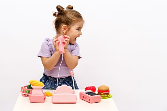 little girl plays shop with toy cash register, scales and plastic fruits and vegetables on white table at home, looks away and smiles, copy space