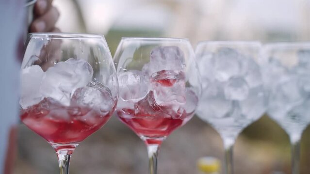 Preparing cool summer bright cocktails. Beautiful transparent glasses with ice cubes stand in row. Bartender from metal measuring cup, jigger pours red drink into bowl glass, close-up.
