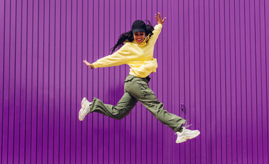 Fototapeta na wymiar Jumping young woman in yellow dress on purple background. Dancer of hip hop and trap music. Concept of freedom, celebration, joy.