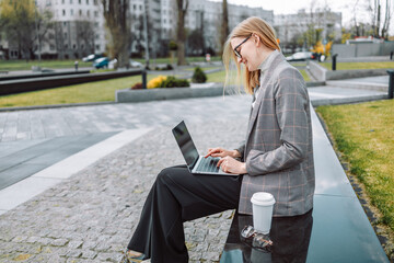 Positive30s business female with coffee sitting in the park and using laptop outdoors, , working remotely, using banking application. 