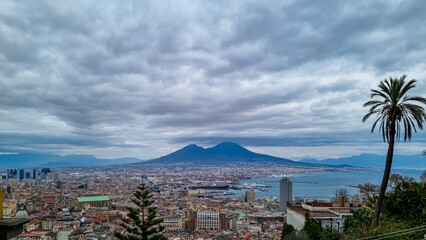 Fototapeta premium Panoramic view from Vomero on volcano Mount Vesuvius in Naples, Campania, Italy, Europe. Ferries in the port of Naples. Clouds are accumulating around the mountains. Palm tree with sea view