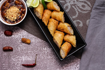 Fried spring rolls in a black plate served with tamarind dipping sauce on the table with black and...