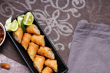 Fried spring rolls in a black plate served with tamarind dipping sauce on the table with black and...