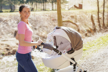 Young woman wearing casual clothes pushing baby carriage stroller on country road, sunny day, blurred trees background