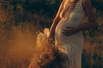 A pregnant woman in a white long dress with a bouquet of dried flowers at sunset time....