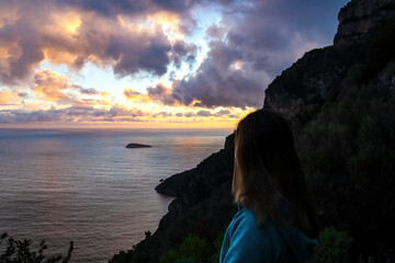 Woman watching the sunset over island of Scoglio Vetara on Mediterranean sea at Amalfi Coast, Campania, Italy, Europe. Golden hour sunlight on surface of Tyrrhenian Sea. Red orange clouds emerging.