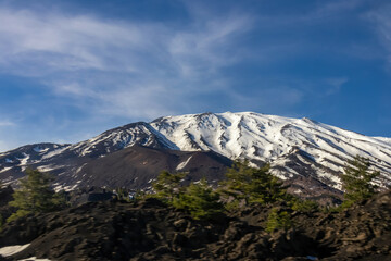 Panoramic view on north eastern flank of volcano mount Etna, in Sicily, Italy, Europe. Summit is covered with snow. Landscape is black brown volcanic sand, bare terrain. Smoke is around the craters