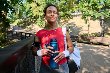 Confident athletic woman in red tight t-shirt and terry wristbands holds bottle with water and...