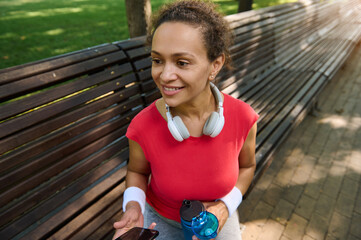 Middle aged African American fit woman, athlete in bright red t-shirt smiles while relaxing sitting...