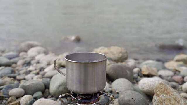 Idyllic Camping Scene. Water in a Metal Mug Boils on a Gas Burner. Hike. Breakfast on the River Bank
