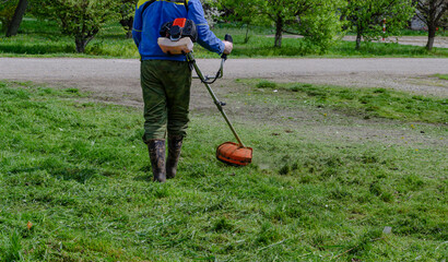 A close-up of a worker in protective clothing, gloves, rubber boots with a gas mower on the front...