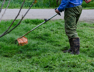 A close-up of a worker in protective clothing, gloves, rubber boots with a gas mower on the front lawn. A man mows grass with dandelions on a warm sunny spring day.