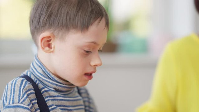 Side view smiling autistic curios boy licking palm choosing colors for painting in school. Portrait of Caucasian child with birth anomaly studying at home with unrecognizable teacher