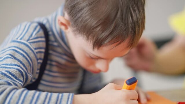 Close-up concentrated autistic Caucasian boy painting with tongue out sitting at table indoors. Focused child with mental illness drawing studying fine art in school. Autism and individuality concept