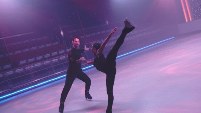 Figure skaters pair on ice rink in neon light skating by camera,man moves backwards holding woman hand,performing arabesque,leg and arm raised parallel high up behind.Slow motion. Camera tracking
