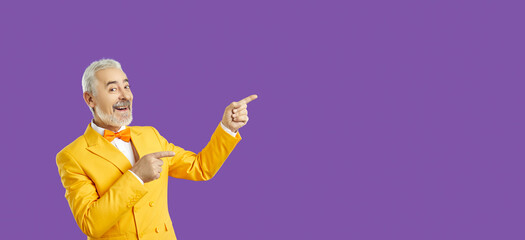 Middle aged man with a beard and grey-haired in a yellow jacket, bow tie and glasses is impressed, looking at camera and pointing away side with his finger. Stands on isolated purple background.