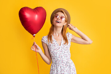 cheerful girl in straw hat and sunglasses hold love heart balloon on yellow background