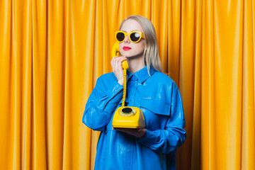 Stylish woman in blue shirt and yellow sunglasses stands with retro dial phone on yellow curtains background