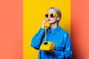 Stylish woman in blue shirt and yellow sunglasses with dial phone on yellow background