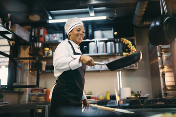 Happy black female chef has fun while preparing food in frying pan at restaurant kitchen. - obrazy, fototapety, plakaty