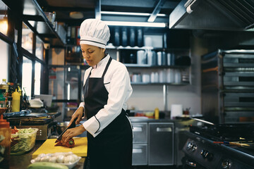 African American female cook cuts vegetables while preparing food in kitchen at restaurant.