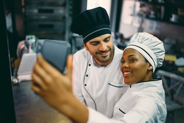 Happy black chef and her male coworker taking selfie with smart phone at work.