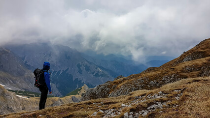 Fototapeta premium Man with backpack on scenic hiking trail with view on cloud covered mountain peaks of the Hochschwab Region in Upper Styria, Austria. Dry alpine meadows in beautiful Alps in Europe. Freedom vibe. Rain