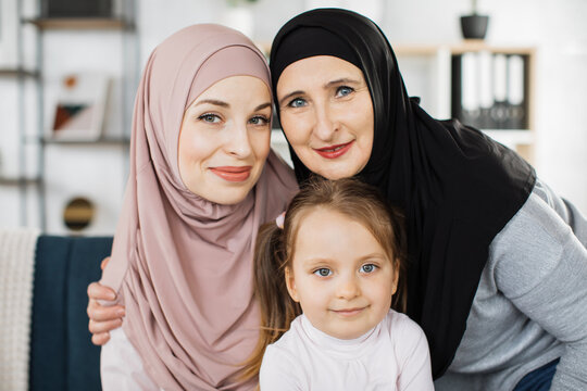 Portrait of three generations of muslim women look at camera posing for family picture, cute little girl hug arabian mom in hijab and granny enjoy time at home, smiling mother daughter and grandmother - Powered by Adobe