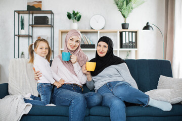 Three muslim female generations family portrait. Cute girl, islamic mom and grandma hold mugs of hot drinks. Mother, grandmother and granddaughter on couch at home, enjoying leisure time together