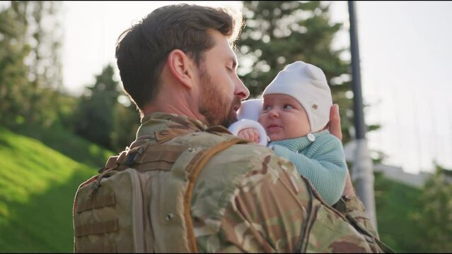 At sunlight father soldier in uniform hugging kissing cute newborn daughter outdoors at sunlight. Stand in the park. Children. Army. Military concept