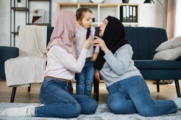 Three generations of Muslim women sitting on their knees on the floor near the sofa in the living...