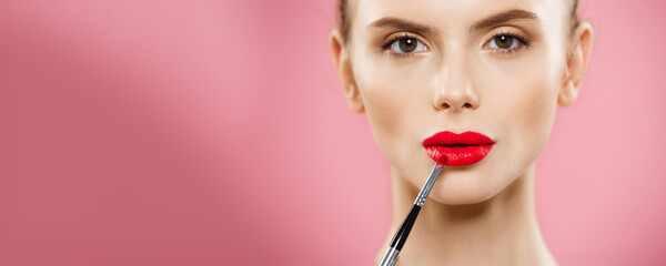 Beauty Concept - Woman applying red lipstick with pink studio background. Beautiful girl makes...