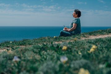 adult woman thoughtfully writes plans diary on a sunny day on a mountain against backdrop of the sea. pensioner sits on the grass barefoot in summer, spring, with a notebook and a pen