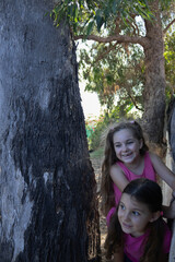 two little girls playing behind a tree with a big smile on their faces 